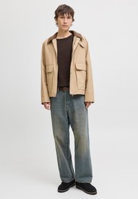 Unge mand iført en beige jakke over en mørkebrun skjorte, løse falmede blå jeans og sorte sko, stående foran en ensfarvet lys baggrund.