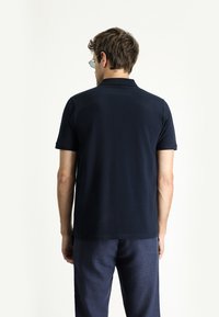 Polo di colore blu navy realizzato in tessuto morbido, con maniche corte e colletto classico. Indossato con pantaloni grigi a trama, visto da dietro.