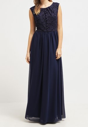 Kobieta ubrana w granatową suknię bez rękawów z koronkowym gorsetem i zwiewną spódnicą z szyfonu, stojąca na tle gładkiego jasnego tła.