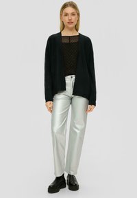 Schwarze offene Strickjacke über einem dunkel gepunkteten Top, kombiniert mit glänzenden silbernen geraden Hosen und schwarzen klobigen Stiefeln.