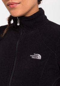 Mustat fleece-takki korkealla kauluksella, etuvetoketjulla ja brodeeratulla logolla rinnassa. Pehmeä tekstiili, yksinkertainen muotoilu ja urheilullinen istuvuus.