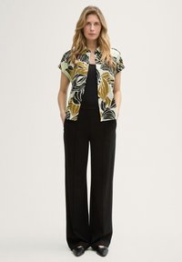 Camicia a maniche corte di colore verde chiaro con motivi di foglie nere e dorate, abbinata a pantaloni neri a vita alta e scarpe nere a punta.