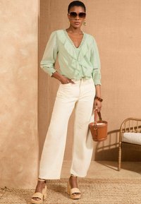 Blusa verde chiaro con frontale a volant, jeans bianchi a gamba larga e sandali con zeppa in paglia. Una borsa a secchiello marrone e occhiali da sole oversize completano il look.