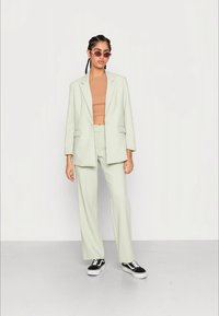 Blazer ajustado verde claro e calças largas combinadas, usadas sobre uma blusa bege. Ténis preto e branco. Acessórios incluem óculos de sol pequenos.