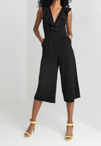 Svart ärmlös jumpsuit med djup V-ringning, volangdetalj fram, breda kapade ben och två sidofickor. Ageras med gula högklackade skor.