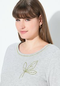Hellgraues T-Shirt mit kurzen Ärmeln, verziert mit grüner Blattstickerei und einem passenden grünen Halsabschluß. Weicher Stoff mit lockerer Passform.