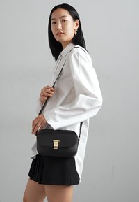 Svart axelremsväska med texturerad yta, guldfärgade detaljer och lockstängning, bärs av en modell i vit skjorta och svart plisserad kjol.