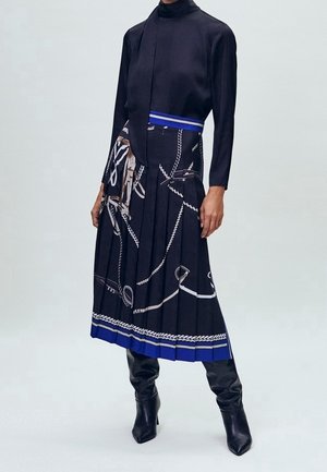 Femme portant un haut noir à manches longues et une jupe plissée noire avec des motifs de corde blanche et une bordure bleue, associée à des bottes noires à talons.