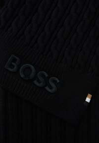 Melna adīta šalle ar pinuma rakstu, izsistā "BOSS" logo un mazu akcenta birku ar strīpām melnā, baltā un zelta krāsā.