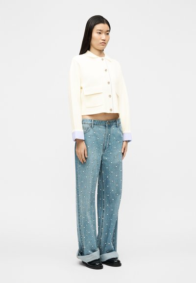 Femme portant un cardigan court crème à boutons dorés et un jean large bleu clair décoré de perles et aux revers retroussés.