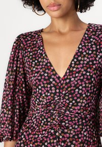 Robe florale avec un décolleté en V profond, ornée de motifs de marguerites roses, rouges et dorées sur un fond noir ; tissu doux avec des manches cloches plissées.