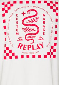 Camiseta blanca con estampado gráfico rojo que presenta una serpiente, hojas y un patrón de ajedrez. El texto incluye "GARAGE PERSONALIZADO" y "REPLAY" establecido en 1981.