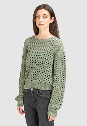 Groene gebreide trui met een open maaspatroon, ronde hals en ribgebreide mouwen. Draagt over zwarte jeans met lange mouwen.