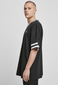 Černé oversized tričko má design s véčkovým výstřihem, krátké rukávy s bílými pruhy a malým logem na hrudi. Hladká, bavlněná textura.