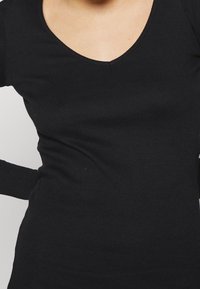 Haut noir côtelé à manches longues avec un col en V. Le tissu semble doux avec une légère texture et une coupe ajustée. Les manches accentuées couvrent les poignets.