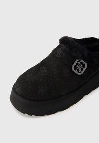 Schwarzer Wildlederschuh mit einem plüschigen Innenfutter, ausgestattet mit einem geprägten Logo-Muster und einem dekorativen Metallelement am Riemen. Flache Gummisohle.