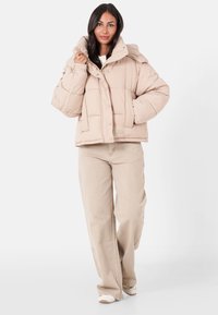 Beige Puffermantel mit hohem Kragen, Druckknöpfen und gestepptem Muster. Kombiniert mit hellbeigen weit geschnittenen Hosen und weißen Schuhen.