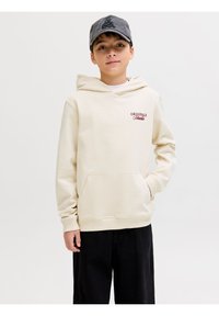 Αγόρι που φοράει μπεζ φούτερ με κουκούλα με το κείμενο "Originals Studio", μαύρο παντελόνι και σκούφο μπέιζμπολ σε σκούρο γκρι, στέκεται με το ένα χέρι στην τσέπη.