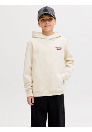 Boy wearing a beige hoodie with "Originals Studio" text, black pants, and a dark gray baseball cap, standing with one hand in pocket.