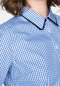 Camisa de cuadros azul y blanca con cuello clásico, botones blancos y ribete negro en el borde del cuello. Tejido de algodón con una textura suave.