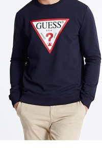 Sudadera azul marino con un logo triangular de GUESS en rojo y blanco, que presenta un signo de interrogación y texto. Ajuste suave y casual con puños de canalé.