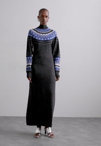 Rochie lungă neagră cu accente tricotate în nuanțe de albastru și alb la guler și manșete, purtată cu pantofi argintii cu vârful deschis. Textură netedă.