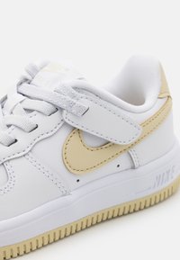 Weiße Ledersneaker mit einem hellbraunen Nike-Swoosh, strukturierten Gummisohlen und Stoffsenkeln. Mit einem Klettverschlussriemen für zusätzlichen Halt.