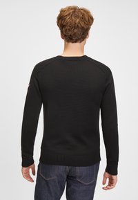 Schwarzer Pullover mit gerippten Bündchen an Ärmeln und Saum, mit einer strukturierten Webart. Gestaltet mit Rundhalsausschnitt und strukturierten Schultern. Kombiniert mit blauen Jeans.
