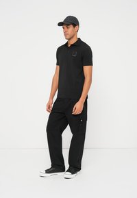 Čierne polokošeľa s krátkymi rukávmi, na ktorej je nenápadné zlaté logo. Kombinované s čiernymi cargo nohavicami a čiernymi teniskami. Na hlave sivá šiltovka.