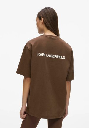 Kvinde med langt hår iført brun oversized t-shirt med "KARL LAGERFELD" logo i hvid på ryggen, stående mod hvid baggrund.