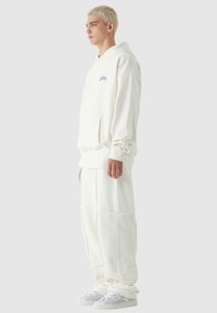Sudadera blanca de forro polar con bolsillo frontal y logo bordado, combinada con pantalones holgados a juego, que presentan detalles de patchwork.