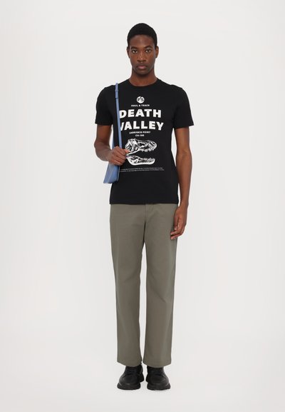 Czarny bawełniany T-shirt z grafiką "DEATH VALLEY", oliwkowe spodnie i czarne buty; trzyma małą jasno-niebieską torbę.