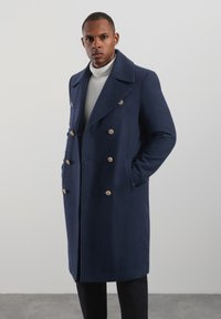 Marineblå uldfrakke med dobbeltspænding, store revers, knapper i guldfarve og frontlommer. Stylet over en grå sweater.