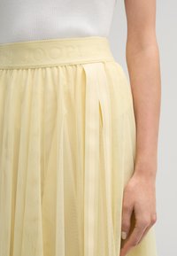 Gelber Netzrock mit einem strukturierten Taillenbund, auf dem "JOOP!" eingeprägt ist. Er hat durchsichtige Lagen und vertikale Bändchenakzente für zusätzliche Details.