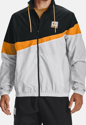 Homme portant une veste coupe-vent à blocs de couleurs noir, orange et blanc, avec une fermeture éclair à l'avant, et un pantalon noir avec des écussons logo.