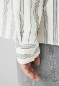 Chemise blanche à fines rayures noires, avec un poignet à fermeture à un bouton et un tissu texturé. Main reposant sur un fond en tissu gris.