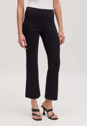 Woman wearing black flared pants and black heeled sandals, standing against a plain neutral background.