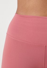 Visok pas, pastelno roza leggingi iz gladkega materiala; imajo ravne šive in prilegajoč dizajn za eleganten silhuet.