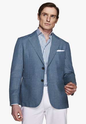 Hombre con un blazer azul sobre una camisa a rayas azules y blancas, con un pañuelo blanco en el bolsillo y pantalones blancos, posando frente a un fondo liso.