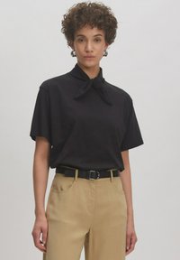 Zwarte korte mouw shirt met een geknoopt kraagdetail, gecombineerd met beige hoog gewaisted broek en een zwarte riem. Glad stoffen textuur.