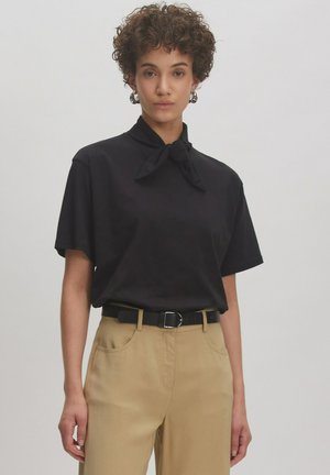 Zwarte korte mouw shirt met een geknoopt kraagdetail, gecombineerd met beige hoog gewaisted broek en een zwarte riem. Glad stoffen textuur.