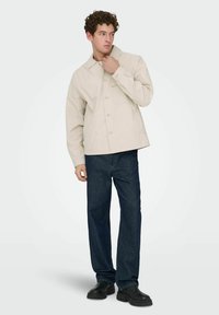 Veste beige à boutons avec col et une poche poitrine unique, associée à un jean à jambes droites bleu foncé et des chaussures noires.