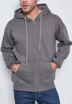 Homme portant un sweat à capuche gris avec fermeture éclair, cordons et poches avant, les mains partiellement dans les poches, associé à un jean bleu.