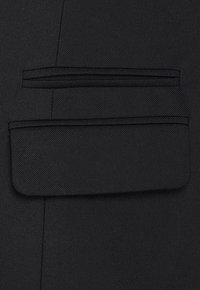 Tessuto nero con finitura testurizzata che presenta una tasca orizzontale e una tasca rettangolare con pattina su un design sartoriale.