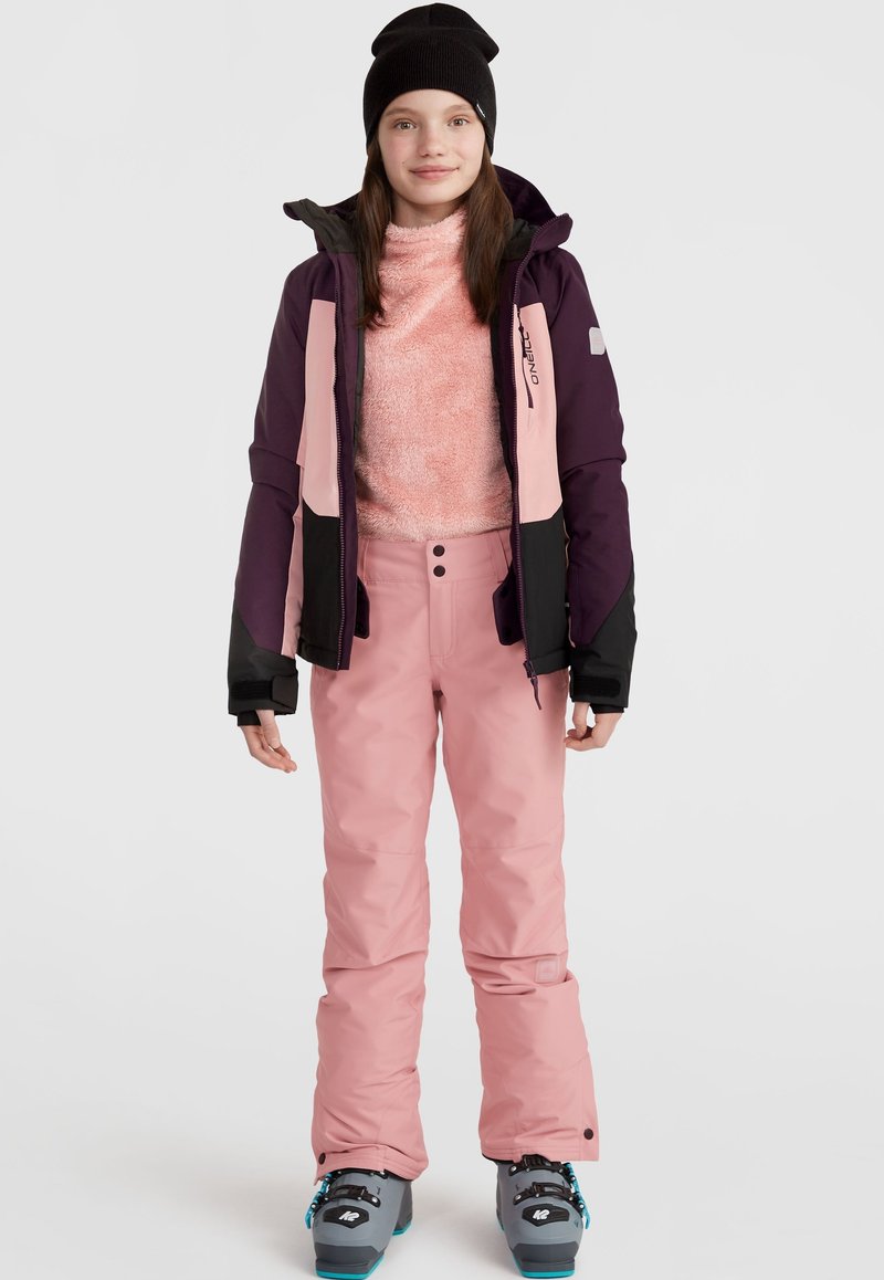 Tenue de ski d'hiver composée d'une veste violette et rose avec des accents noirs, d'un t-shirt en polaire rose et d'un pantalon de ski rose. Portée avec des bottes noires.