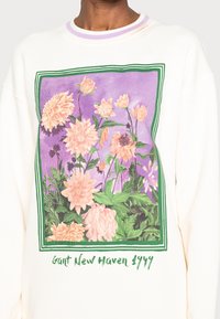Sudadera blanca con un gráfico floral en rosa y morado dentro de un marco verde, con el texto "Gant New Haven 1949" en la parte inferior.