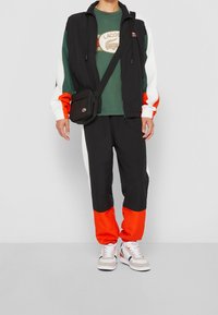 Herrträningsdräkt med en svart jacka med vita och orange detaljer, och svarta byxor med orange muddar. Kombinerad med en grön t-shirt och sneakers.