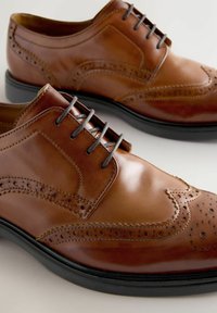 Un par de zapatos de vestir brogue de cuero marrón pulido con suelas negras y cordones grises sobre un fondo claro.