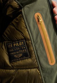 Olijfgroene gewatteerde binnenkant van een jacket met een gouden ritszak. Bevat een zwart label met de tekst "ICE PILOT" in witte letters en logo.