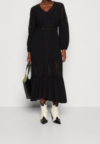 Abito midi nero con dettagli in pizzo, maniche lunghe, chiusura a bottoni e gonna a strati. Abbinato a stivali cowboy a punta bianchi e neri e una borsa nera.